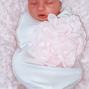May include: A white knotted gown with a large pink flower on the front. The gown is worn by a baby wearing a white hat with a pink flower on top.