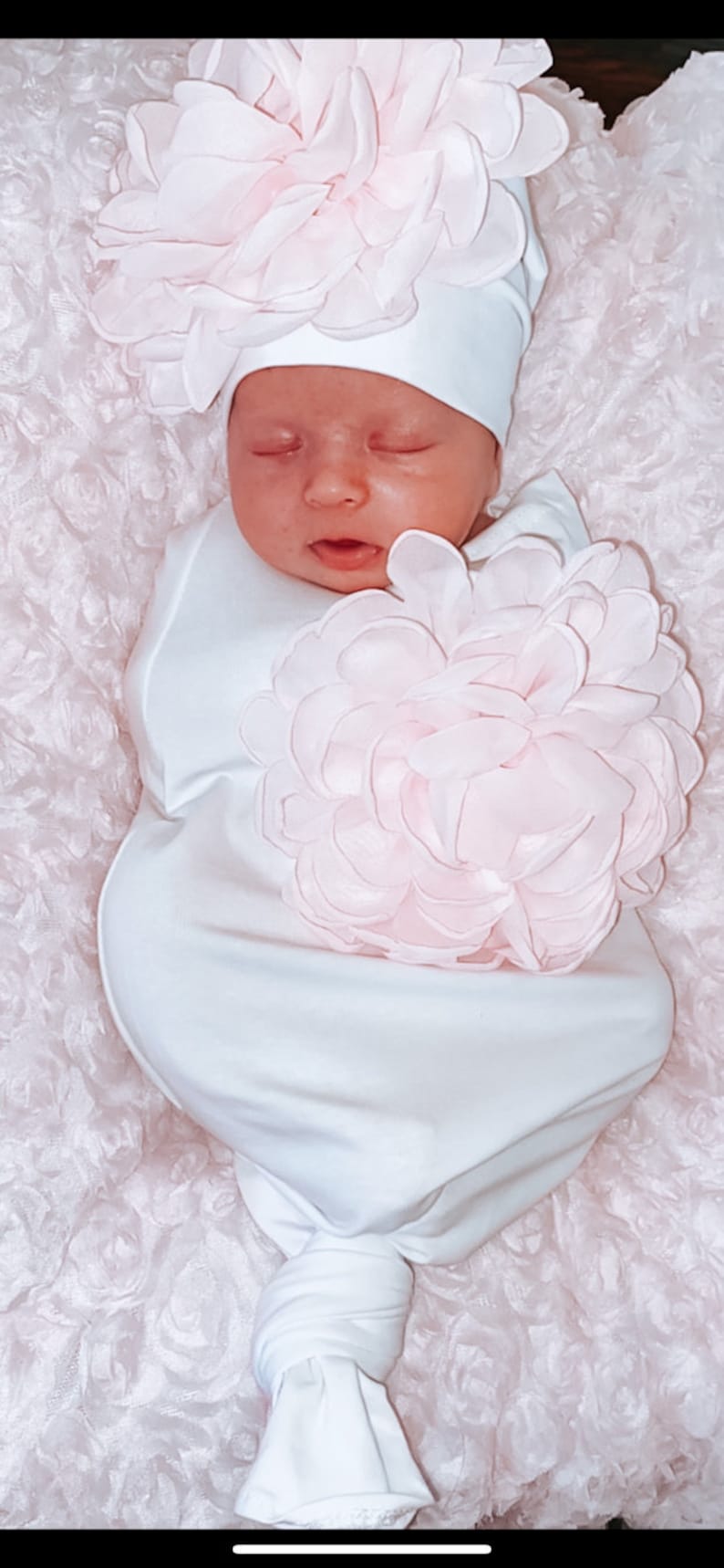 May include: A white knotted gown with a large pink flower on the front. The gown is worn by a baby wearing a white hat with a pink flower on top.