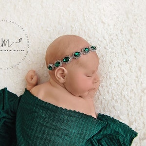 K&ouml;nnte beinhalten: Ein neugeborenes Baby, das in ein gr&uuml;nes Swaddle gewickelt ist und ein silbernes Stirnband mit smaragdgr&uuml;nen Edelsteinen tr&auml;gt. Das Baby schl&auml;ft friedlich auf einem wei&szlig;en Hintergrund.