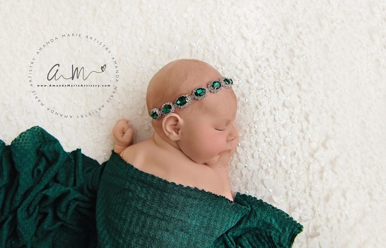 K&ouml;nnte beinhalten: Ein neugeborenes Baby, das in ein gr&uuml;nes Swaddle gewickelt ist und ein silbernes Stirnband mit smaragdgr&uuml;nen Edelsteinen tr&auml;gt. Das Baby schl&auml;ft friedlich auf einem wei&szlig;en Hintergrund.