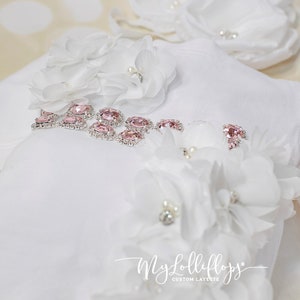 Pode incluir: Macac&atilde;o branco para beb&ecirc; com detalhes rosa e prateados. O macac&atilde;o &eacute; decorado com flores de tecido branco e strass. O texto "My Lolliflops Custom Layette" est&aacute; impresso no macac&atilde;o.