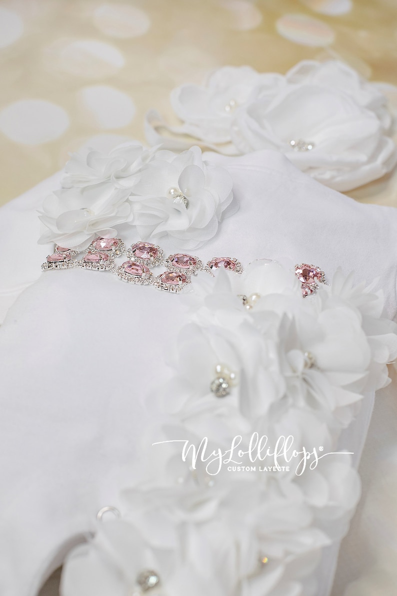Pode incluir: Macac&atilde;o branco para beb&ecirc; com detalhes rosa e prateados. O macac&atilde;o &eacute; decorado com flores de tecido branco e strass. O texto "My Lolliflops Custom Layette" est&aacute; impresso no macac&atilde;o.