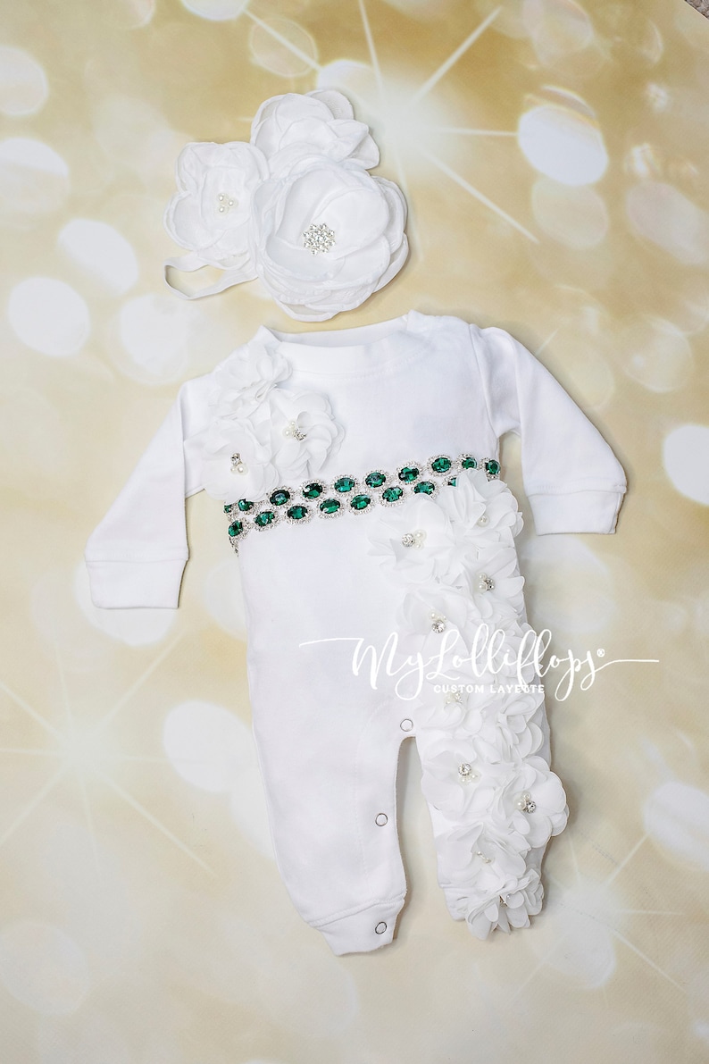 Pode incluir: Macac&atilde;o branco para beb&ecirc; com um cinto de joias verdes e enfeites de flores brancas. O macac&atilde;o tem bot&otilde;es de press&atilde;o na parte inferior e &eacute; decorado com flores brancas. O macac&atilde;o est&aacute; sobre um fundo dourado. O texto "My Lollifloss Custom Layette" est&aacute; no macac&atilde;o.