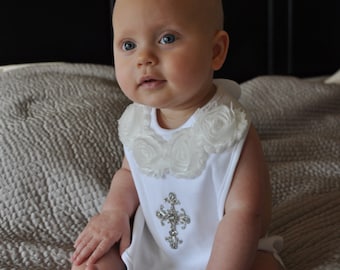 Peut inclure: Un bébé portant une robe blanche avec une bordure de fleurs blanches et une croix en strass sur le devant. Le bébé est assis sur une couverture texturée grise.