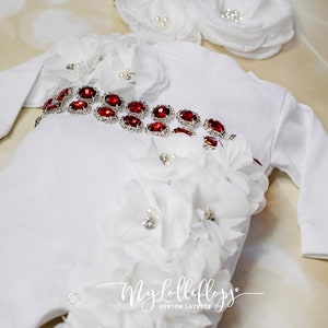 Pode incluir: Um macac&atilde;o branco para beb&ecirc; com um cinto vermelho adornado com joias e flores brancas. O macac&atilde;o tem mangas compridas e um fecho de press&atilde;o na parte inferior. O texto "My Lolliflops Custom Layette" est&aacute; impresso na parte inferior do macac&atilde;o.