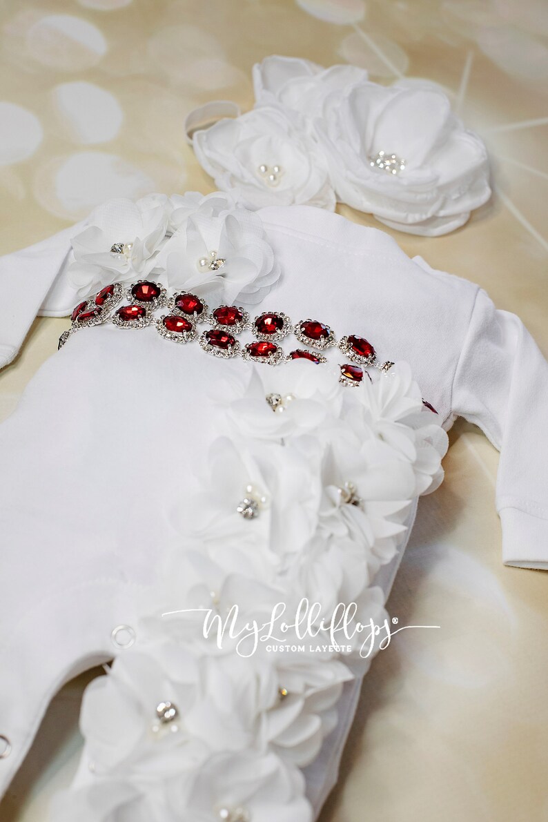 Pode incluir: Um macac&atilde;o branco para beb&ecirc; com um cinto vermelho adornado com joias e flores brancas. O macac&atilde;o tem mangas compridas e um fecho de press&atilde;o na parte inferior. O texto "My Lolliflops Custom Layette" est&aacute; impresso na parte inferior do macac&atilde;o.