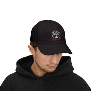 Könnte beinhalten: Eine schwarze Baseballkappe mit einer kreisförmigen Grafik auf der Vorderseite. Das Design zeigt einen weißen Totenkopf mit Feuerwehrhelm, gekreuzte Äxte und den Schriftzug FIRST ALARM FATHERHOOD in weißer und roter Schrift auf dunklem Grund.