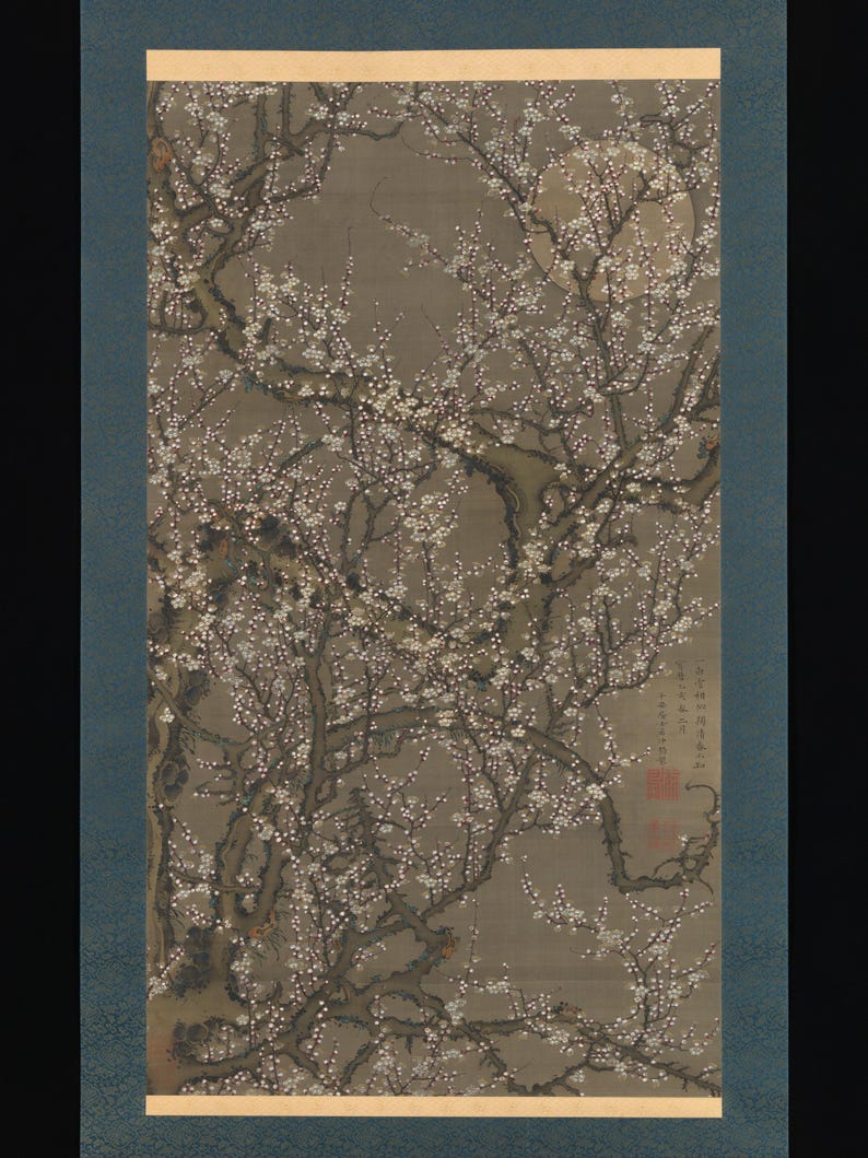 May include: A vertical hanging scroll painting on silk depicting a gnarled plum tree in full bloom with white blossoms against a muted gray background. A pale, circular moon is visible in the upper right. The artwork includes black ink calligraphy and two red seal stamps in the lower right corner.