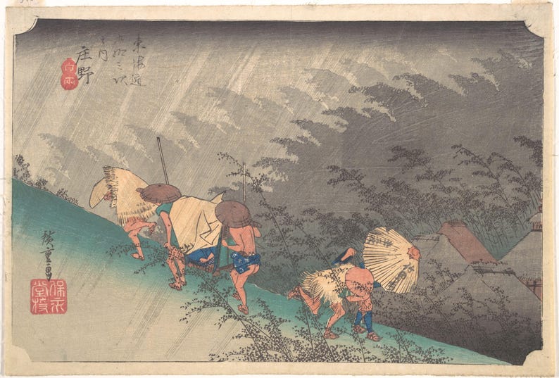 May include: A Japanese woodblock print depicting travelers walking along a path during a rainstorm. The figures wear straw capes and hats, with one person holding a paper umbrella. The scene features muted tones of green, brown, and grey.