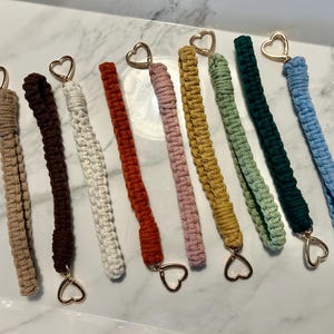 May include: A collection of eight macrame wristlet keychains in various colors including beige, dark brown, white, rust, dusty pink, mustard yellow, sage green, dark green, and light blue, each featuring a gold-toned metal heart-shaped clasp.