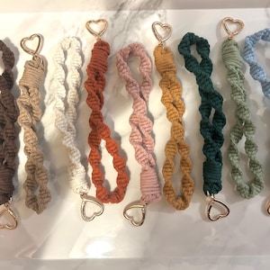 May include: A collection of nine macrame wristlet keychains in various colors including dark brown, tan, cream, rust, dusty pink, mustard yellow, dark green, sage green, and light blue, each featuring a gold-toned metal heart-shaped clasp.
