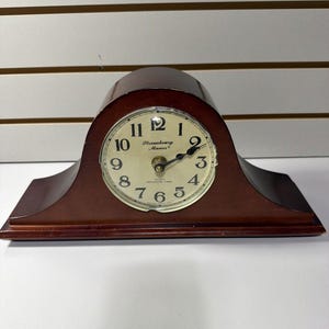 May include: A dark brown wooden mantel clock with an arched top and a rectangular base. The clock face is off-white with black Arabic numerals, black hands, and the text Strasbourg Manor and Quartz Westminster Chime printed in black.