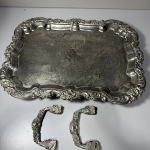 May include: A rectangular silver-toned metal serving tray with an ornate, scalloped edge featuring embossed floral details. The tray has a tarnished surface with visible wear. Two matching, detached metal handles with intricate scrollwork.