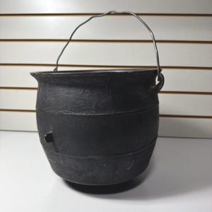 May include: A black cast iron cauldron with a rounded body, a wide rim, and a thin metal wire handle attached to two side lugs. The exterior surface features a textured finish with horizontal ridges and a small protruding tab on the side.