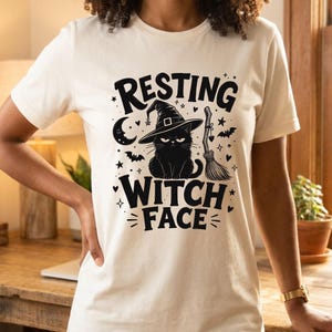 May include: A cream-colored short-sleeve t-shirt featuring a black graphic print of a cat wearing a witch hat. The text reads "RESTING WITCH FACE" in bold black letters, surrounded by illustrations of a crescent moon, stars, bats, and a broom.