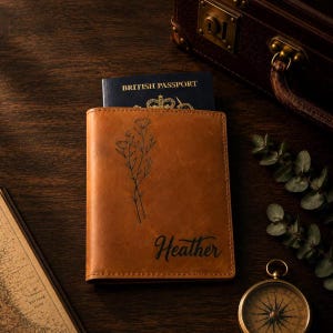 Pode incluir: Um porta-passaporte de couro castanho com uma ilustração de linhas pretas de flores silvestres e o nome Heather gravado em escrita cursiva preta. Um passaporte britânico azul escuro está inserido com o texto British Passport visível.
