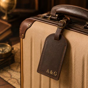 Op de afbeelding: Een donkerbruine lederen bagagelabel met goudkleurige letters A & G, bevestigd aan een beige koffer met donkerbruine lederen afwerking en een handvat. Het label is vastgemaakt met een dunne lederen riem en een kleine koperen gesp.