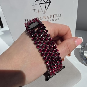 May include: A watch band featuring four rows of deep red circular crystals set in a dark metal frame. The band includes a rectangular black clasp and a connector piece. The background displays a white card with the text HANDCRAFTED BANDS.