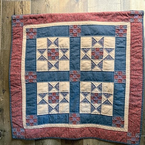May include: A square quilted wall hanging featuring a four-block star pattern. The design incorporates cream, dusty blue, and muted red floral fabrics. The border is a deep red floral print with small blue and purple square accents at the corners.