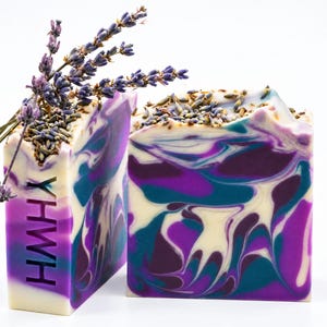 May include: Two rectangular bars of soap featuring a marbled swirl pattern in shades of purple, teal, and white. The tops are decorated with dried lavender buds, and one side of a bar is stamped with the letters YHWH in a vertical orientation.