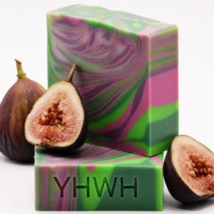May include: Two rectangular bars of soap with a marbled pattern of purple, pink, and green swirls. The front bar is stamped with the letters YHWH. The soap is arranged with one whole fig and two halved figs showing the seeded interior flesh.
