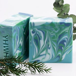 May include: Two rectangular bars of soap featuring a marbled pattern of blue, green, and white swirls. One bar is positioned upright, displaying the letters YHWH embossed vertically into its side. A sprig of green leaves is placed nearby.