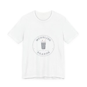 May include: A white short-sleeved t-shirt featuring a circular graphic on the chest. The design includes a drawing of a drink in a glass with a straw and a cherry, surrounded by the text MUDSLIDE SEASON and CAPE COD, MASS in a dark grey font.