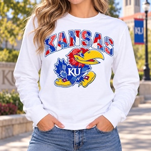 May include: A white long-sleeve sweatshirt featuring the word KANSAS in a red, white, and blue patchwork pattern. Below the text is a graphic of a Jayhawk mascot in blue and red with a yellow beak, also featuring a patchwork design and the letters KU.