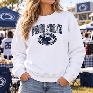 May include: A white crewneck sweatshirt featuring the text PENN STATE in a dark blue, patterned font arched above an oval graphic containing a dark blue Nittany Lion logo. The sweatshirt has long sleeves and a ribbed collar, cuffs, and hem.