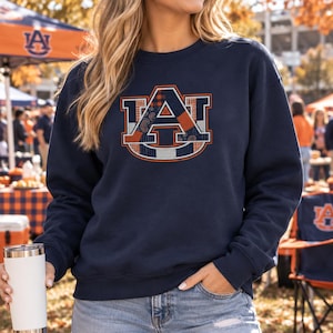 May include: A navy blue long-sleeve sweatshirt featuring a large Auburn University AU logo on the chest. The logo is filled with a patchwork pattern of orange, navy, and white colors, including plaid and paisley designs, outlined in white and orange.