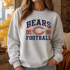 May include: A light gray crewneck sweatshirt featuring a distressed navy blue and orange graphic print that reads BEARS EST. 1920 FOOTBALL with a large orange letter C in the center. The long-sleeved pullover has a ribbed collar, cuffs, and hem.