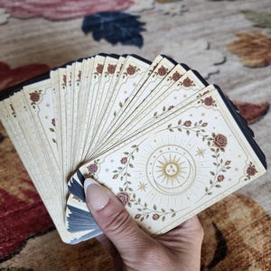 May include: A deck of tarot cards fanned out, featuring a cream-colored background with a central gold sun and moon illustration. The cards are decorated with a border of red roses, green leaves, and small gold stars, with black edges on each card.
