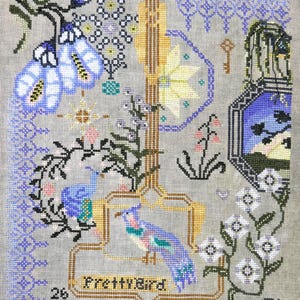 May include: Cross-stitch embroidery featuring a colorful bird within a gold frame, with the text "Pretty Bird." The design includes floral and geometric patterns in shades of blue, green, pink, and white on a gray background. A key and birdcage are also featured.