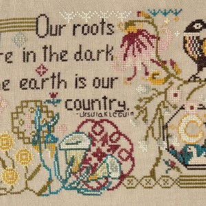 May include: Cross-stitch artwork with the quote "Our roots are in the dark. The earth is our country." by Ursula K. Le Guin. The design features floral and bird motifs in various colors, including pink, yellow, and teal, on a beige background.