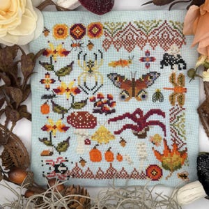 Könnte beinhalten: Ein hellblaues Stoffquadrat mit einem Kreuzstichmuster aus verschiedenen herbstlichen Motiven, darunter eine Spinne, ein Schmetterling, eine Libelle, Pilze, Kürbisse, ein Ahornblatt und geometrische Bordüren in Orange-, Braun- und Gelbtönen.