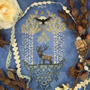 Könnte beinhalten: Eine Kreuzstichstickerei auf blauem Stoff mit einem zentralen Elch, der zwischen zwei Birken steht, und einem Vogel, der über einem Vollmond fliegt. Das Design enthält komplexe geometrische Muster an der Basis und gelbe Laubakzente.