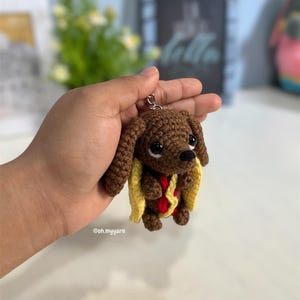 Puede incluir: Un llavero de perro salchicha tejido a ganchillo en color marrón, disfrazado de perrito caliente con un pan amarillo y detalles de mostaza roja. El pequeño peluche tiene ojos de plástico negro y nariz negra, con una anilla metálica plateada.
