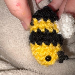 Peut inclure: Un petit jouet abeille crocheté à la main avec des rayures horizontales alternées de fil jaune vif et noir. L'abeille possède un œil en perle de plastique noir visible et une petite aile crochetée blanche texturée sur le côté.