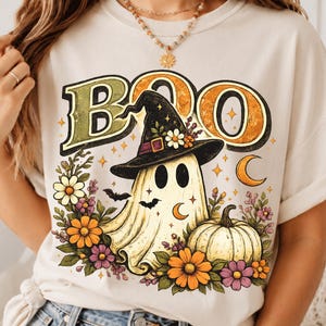 Könnte beinhalten: Ein cremefarbenes T-Shirt mit einem Grafikdruck eines Geistes, der einen Hexenhut trägt, umgeben von orangefarbenen und violetten Blumen, einem Kürbis und kleinen Fledermäusen. Das Wort BOO ist in großen, stilisierten Buchstaben aufgedruckt.