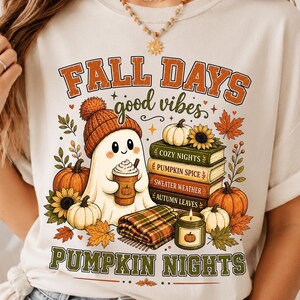Könnte beinhalten: Ein cremefarbenes T-Shirt mit einem Grafikdruck mit dem Text FALL DAYS und PUMPKIN NIGHTS. Das Design zeigt einen Geist mit Strickmütze, der einen Kaffeebecher hält, einen Stapel Bücher mit der Aufschrift COZY NIGHTS, PUMPKIN SPICE, SWEATER WEATHER und AUTUMN LEAVES, Kürbisse, Sonnenblumen, eine karierte Decke und eine Kerze.