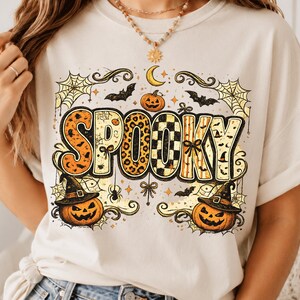 Könnte beinhalten: Ein cremefarbenes T-Shirt mit einem großen Grafikdruck, der das Wort SPOOKY in fetten, gemusterten Buchstaben zeigt. Das Design umfasst orangefarbene Kürbisse mit Hexenhüten, schwarze Fledermäuse, Spinnennetze, eine Mondsichel und Spinnen.