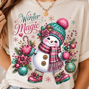 Könnte beinhalten: Ein cremefarbenes T-Shirt mit einer Grafik eines Schneemanns, der eine türkisfarbene Strickmütze mit rosa Bommel, einen karierten Schal und rosa Stiefel trägt. Der Schneemann hält Zuckerstangen und Weihnachtskugeln, umgeben von Blumen und Schneeflocken mit dem Text Winter Magic.