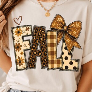 Könnte beinhalten: Ein cremefarbenes T-Shirt mit dem Wort FALL in großen Blockbuchstaben. Das F hat ein Blumenmuster, das A ein Leopardenmuster, das L ein braunes Karomuster und das zweite L schwarze Punkte. Eine große braune Blumenschleife liegt auf den Buchstaben.