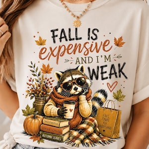 Könnte beinhalten: Ein cremefarbenes T-Shirt mit einer Grafik eines Waschbären mit Sonnenbrille und Schal, der einen Kaffeebecher hält. Der Text lautet FALL IS expensive AND I'M WEAK. Darunter befinden sich Bücher mit der Aufschrift SWEATER WEATHER, PUMPKIN EVERYTHING und AUTUMN LEAVES.