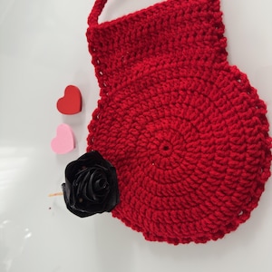 Puede incluir: Una pieza circular tejida a ganchillo en color rojo con un asa superior, acompañada de un pequeño corazón rojo, un pequeño corazón rosa y una rosa artificial negra. El objeto tejido presenta un patrón de puntos concéntricos y borde festoneado.