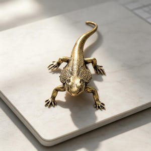 May include: A metallic gold-toned lizard figurine featuring a textured, scaly surface across its back and head. The lizard is depicted in a crawling position with four legs extended outward and a long, curved tail extending behind its body.