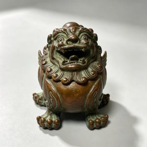 May include: A bronze-toned metal figurine of a mythical lion creature with a wide, open mouth, prominent teeth, and a detailed mane. The sculpture features a weathered patina with dark brown and oxidized green highlights across its textured surface.