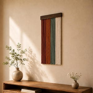 May include: A wall hanging featuring vertical strands of yarn in shades of deep red, burnt orange, sage green, and cream, suspended from a dark brown wooden bar. The textile art is displayed against a plain wall above a wooden console table.