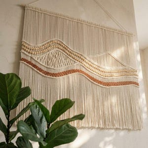 May include: A large macrame wall hanging made of off-white cotton cord, featuring a mountain-inspired landscape design with rows of natural wooden beads and a horizontal band of terracotta-colored woven cord, suspended from a wooden dowel.