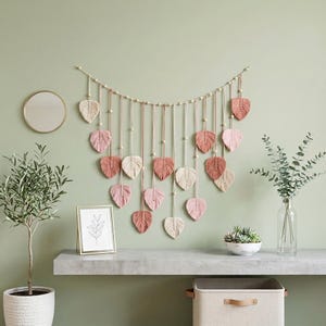 May include: A macrame wall hanging featuring a horizontal cord draped with wooden beads and multiple dangling leaf-shaped fiber ornaments in shades of cream, dusty pink, and terracotta, arranged in a symmetrical, cascading V-shaped pattern.